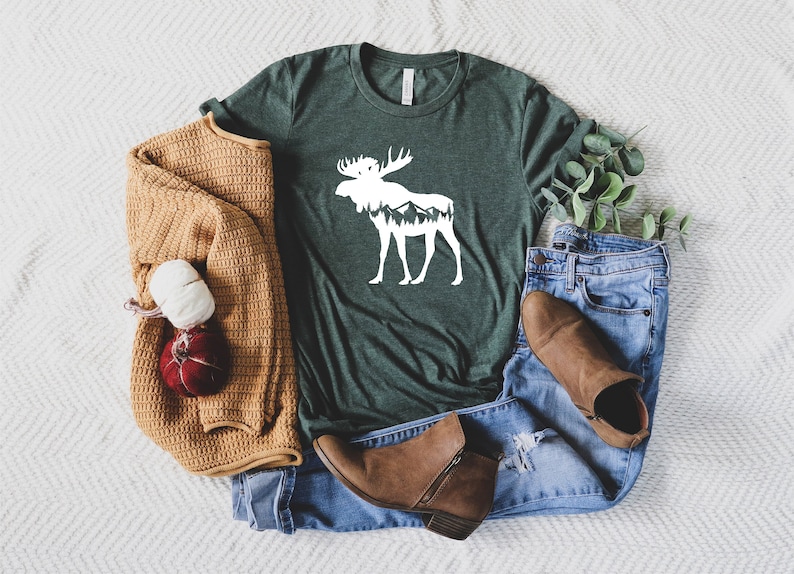 May include: A dark green t-shirt with a white moose graphic, mountains inside the moose silhouette, and a crew neck. The shirt is paired with blue jeans, brown boots, and a tan sweater. A decorative bundle of yarn and a red ornament are also included.