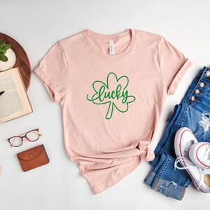 May include: A heather peach t-shirt with a green shamrock and the word "lucky" written in cursive. The shirt is laid out on a white surface with a pair of glasses, a book, a potted plant, a wallet, and a pair of jeans and white sneakers.