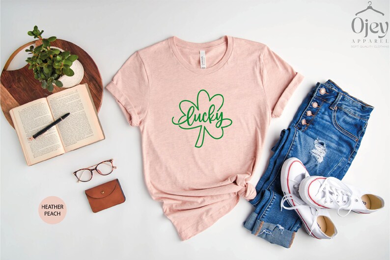 May include: A heather peach t-shirt with a green shamrock and the word "lucky" written in cursive. The shirt is laid out on a white surface with a pair of glasses, a book, a potted plant, a wallet, and a pair of jeans and white sneakers.