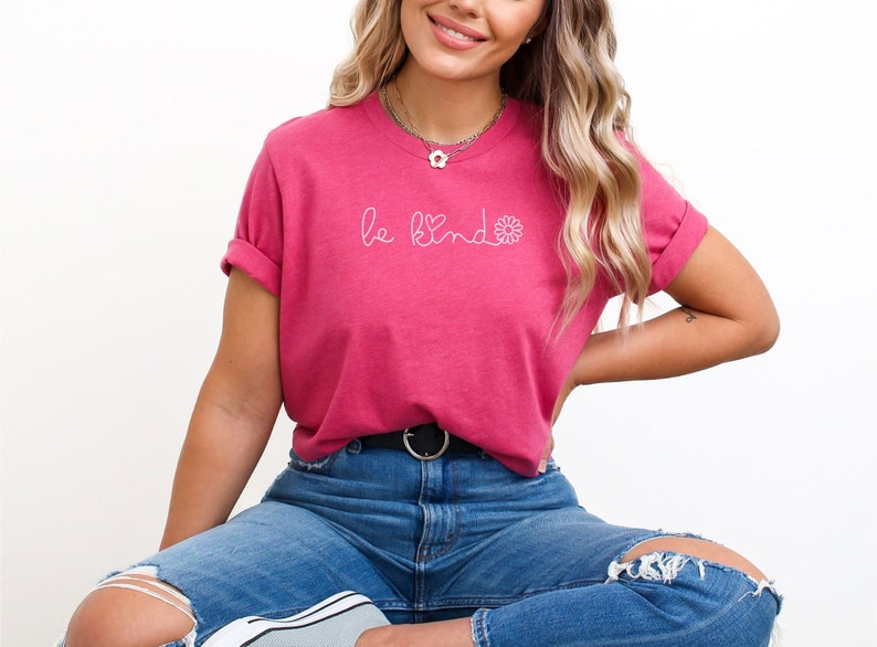 Puede incluir: Una camiseta rosa con el texto "be kind" en blanco con un peque&ntilde;o coraz&oacute;n y un dise&ntilde;o de flor.