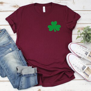 May include: A maroon t-shirt with a green shamrock design. The shirt is paired with blue jeans and white sneakers.