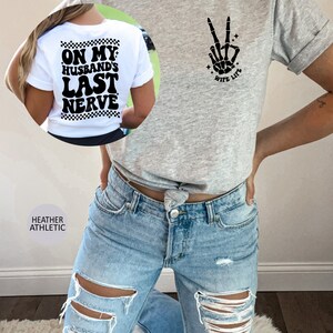 May include: A heather grey t-shirt with a black skeleton hand making a peace sign and the text "Wife Life" on the front. The back of the shirt has the text "On My Husband's Last Nerve" in black with a checkered pattern.