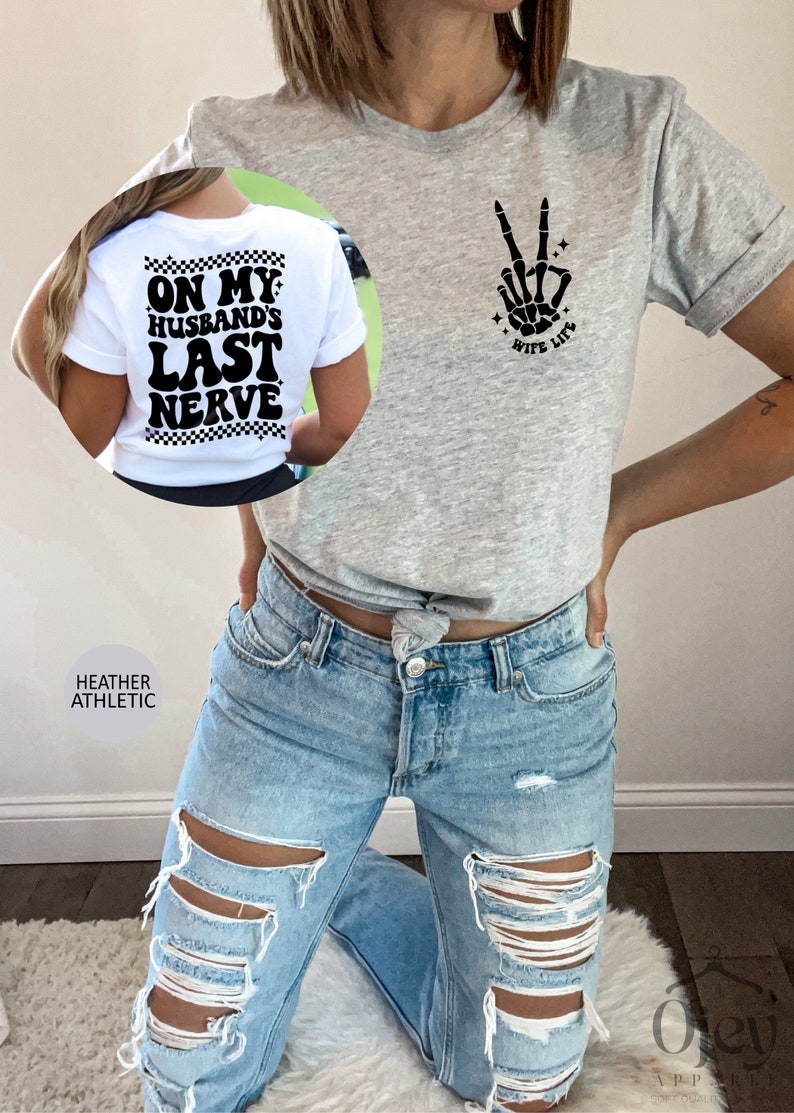 May include: A heather grey t-shirt with a black skeleton hand making a peace sign and the text "Wife Life" on the front. The back of the shirt has the text "On My Husband's Last Nerve" in black with a checkered pattern.