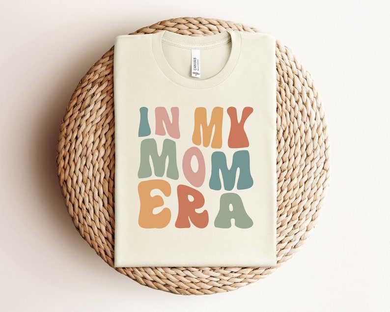 May include: A beige t-shirt with the text "In My Mom Era" in colorful, retro-style lettering.