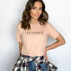 Pu&ograve; includere: Una donna indossa una maglietta rosa chiaro con la scritta "FEYONC&Eacute;" stampata sul davanti. Indossa una camicia a quadri annodata in vita e jeans blu.