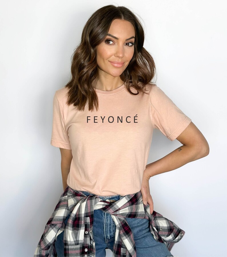 Pu&ograve; includere: Una donna indossa una maglietta rosa chiaro con la scritta "FEYONC&Eacute;" stampata sul davanti. Indossa una camicia a quadri annodata in vita e jeans blu.