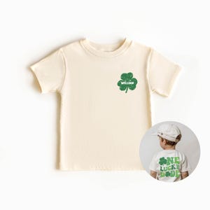 K&ouml;nnte beinhalten: Ein cremefarbenes T-Shirt mit kurzen &Auml;rmeln, auf dem vorne ein gr&uuml;ner vierbl&auml;ttriger Kleeblatt und der Name "William" aufgedruckt sind. Auf der R&uuml;ckseite des Shirts befindet sich ein gr&uuml;ner vierbl&auml;ttriger Kleeblatt und der Text "One Lucky Dude".