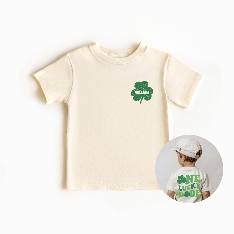 K&ouml;nnte beinhalten: Ein cremefarbenes T-Shirt mit kurzen &Auml;rmeln, auf dem vorne ein gr&uuml;ner vierbl&auml;ttriger Kleeblatt und der Name "William" aufgedruckt sind. Auf der R&uuml;ckseite des Shirts befindet sich ein gr&uuml;ner vierbl&auml;ttriger Kleeblatt und der Text "One Lucky Dude".