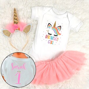 May include: A white onesie with a rainbow unicorn graphic and the text "Birthday Girl". A pink tulle skirt and a gold unicorn headband with pink tulle ears complete the outfit. The onesie also has the name "Sarah" and the number "1" on the back.