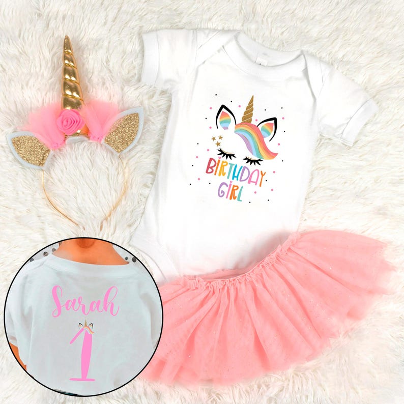 May include: A white onesie with a rainbow unicorn graphic and the text "Birthday Girl". A pink tulle skirt and a gold unicorn headband with pink tulle ears complete the outfit. The onesie also has the name "Sarah" and the number "1" on the back.