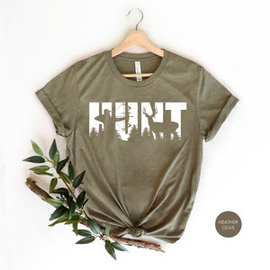 May include: A green t-shirt with the word "HUNT" in white text. The letters are outlined with a silhouette of a hunter with a bow and arrow and a deer.