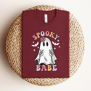 May include: A maroon t-shirt with a white ghost wearing a pink flower. The ghost has its arms raised in a peace sign. The text "Spooky Babe" is printed in pink, orange, and yellow letters.