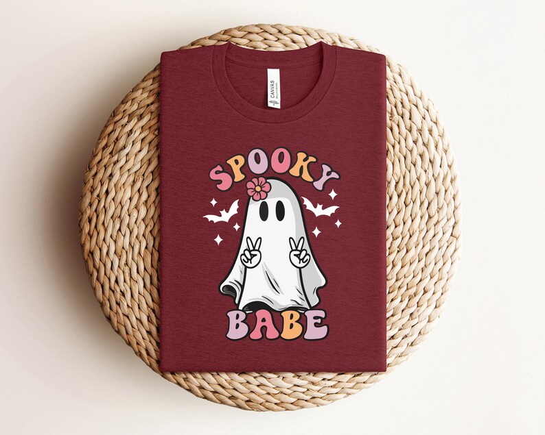 May include: A maroon t-shirt with a white ghost wearing a pink flower. The ghost has its arms raised in a peace sign. The text "Spooky Babe" is printed in pink, orange, and yellow letters.