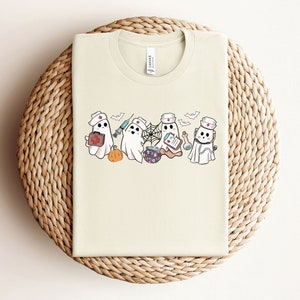 May include: A cream-coloured t-shirt featuring a graphic of four cartoon ghosts dressed as nurses. The ghosts are holding medical supplies such as a syringe, a first aid kit, a potion, and a clipboard.