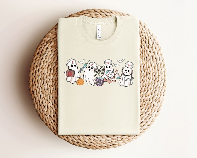 May include: A cream-coloured t-shirt featuring a graphic of four cartoon ghosts dressed as nurses. The ghosts are holding medical supplies such as a syringe, a first aid kit, a potion, and a clipboard.