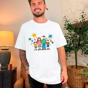 Peut inclure: T-shirt blanc avec une illustration color&eacute;e d'une famille avec un soleil, des c&oelig;urs et des papillons. L'illustration est dessin&eacute;e dans un style enfantin.