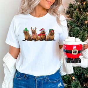 May include: A white t-shirt with a graphic of four seals wearing Christmas hats and decorations. The seals are wearing a Santa hat, a reindeer hat, a Christmas tree hat, and a green elf hat. The seals are wearing string lights around their necks.