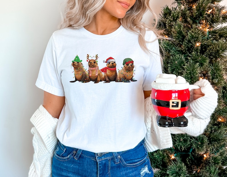 May include: A white t-shirt with a graphic of four seals wearing Christmas hats and decorations. The seals are wearing a Santa hat, a reindeer hat, a Christmas tree hat, and a green elf hat. The seals are wearing string lights around their necks.
