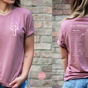 May include: A heather mauve t-shirt with a white cross and the text "Dear Person Behind Me" on the back. The text includes a list of phrases with "You Say" and "God Says" headings.