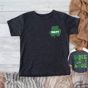 K&ouml;nnte beinhalten: Ein dunkelgraues T-Shirt mit einem gr&uuml;nen Kleeblatt und dem Namen "MATT" darauf gedruckt. Das Shirt ist in Kindergr&ouml;&szlig;e und hat einen Used-Look.