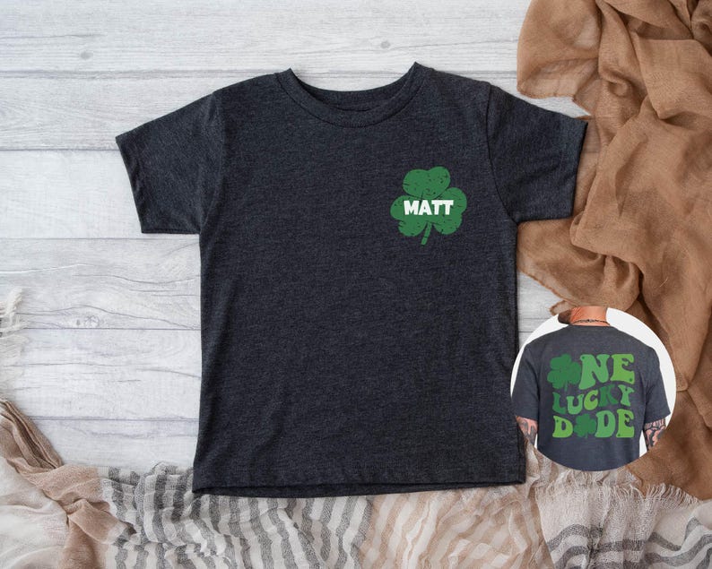 K&ouml;nnte beinhalten: Ein dunkelgraues T-Shirt mit einem gr&uuml;nen Kleeblatt und dem Namen "MATT" darauf gedruckt. Das Shirt ist in Kindergr&ouml;&szlig;e und hat einen Used-Look.