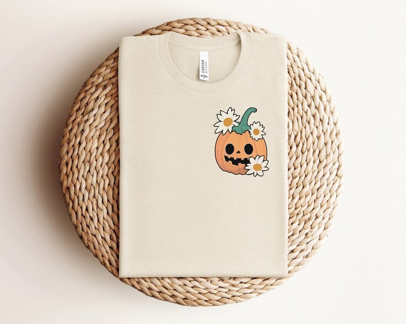 May include: A beige t-shirt with a cartoon pumpkin with a smiling face and a green stem. The pumpkin is surrounded by white daisies.