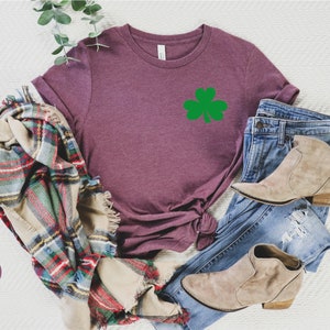 May include: A heather maroon t-shirt with a green shamrock graphic. The shirt is laid out on a white surface with a plaid scarf, blue jeans, and brown ankle boots.