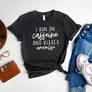K&ouml;nnte beinhalten: Heather dunkelgraues T-Shirt mit dem Text "I run on caffeine and kisses #momlife" in Wei&szlig;. Das Shirt ist auf einem wei&szlig;en Hintergrund mit einer Jeans, einer Handtasche und einem Paar wei&szlig;en Turnschuhen abgebildet.