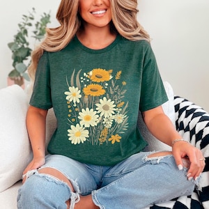 K&ouml;nnte beinhalten: Ein gr&uuml;nes T-Shirt mit einem Blumendruck aus gelben und wei&szlig;en G&auml;nsebl&uuml;mchen. Die G&auml;nsebl&uuml;mchen sind in einem Strau&szlig; mit gr&uuml;nen Bl&auml;ttern und Stielen angeordnet.