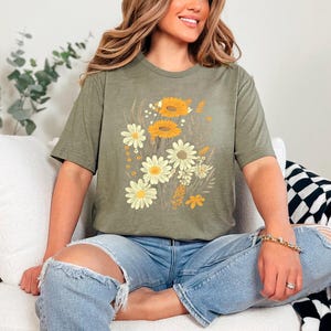 K&ouml;nnte beinhalten: Eine Frau tr&auml;gt ein gr&uuml;nes T-Shirt mit einem Blumendruck mit gelben und wei&szlig;en Blumen. Sie sitzt auf einem wei&szlig;en Sofa mit einer schwarz-wei&szlig;en karierten Wolldecke.
