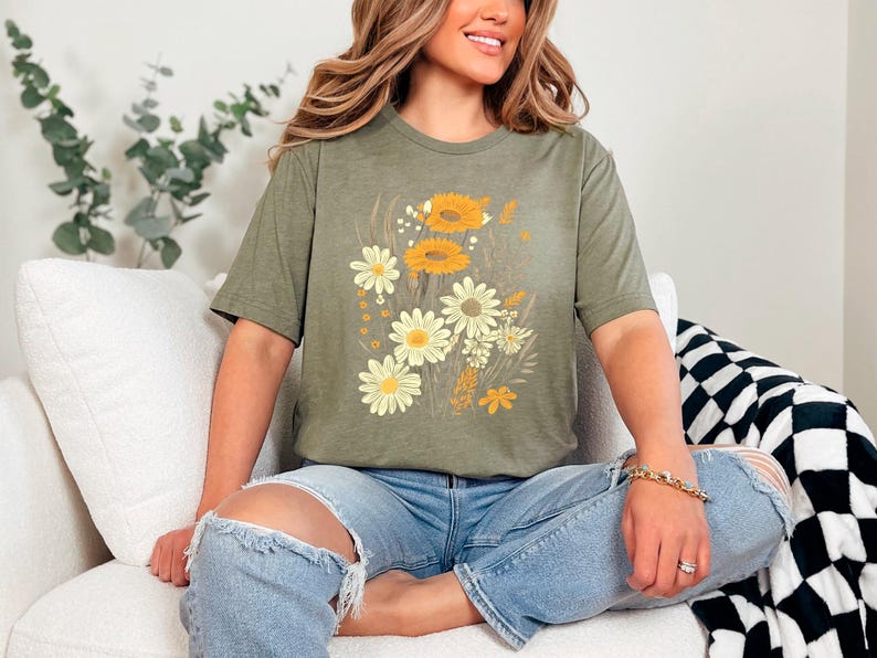 K&ouml;nnte beinhalten: Eine Frau tr&auml;gt ein gr&uuml;nes T-Shirt mit einem Blumendruck mit gelben und wei&szlig;en Blumen. Sie sitzt auf einem wei&szlig;en Sofa mit einer schwarz-wei&szlig;en karierten Wolldecke.