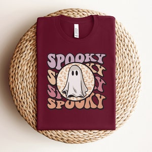 Puede incluir: Una camiseta color granate con un gr&aacute;fico retro de un fantasma en un c&iacute;rculo de estampado de leopardo. El texto "Spooky" est&aacute; escrito con una fuente ondulada alrededor del c&iacute;rculo.