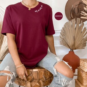 K&ouml;nnte beinhalten: Ein maroonfarbenes T-Shirt mit dem Wort "mama" auf der Vorderseite gestickt. Das Shirt wird von einer Person getragen, die auf einem Holzhocker sitzt.