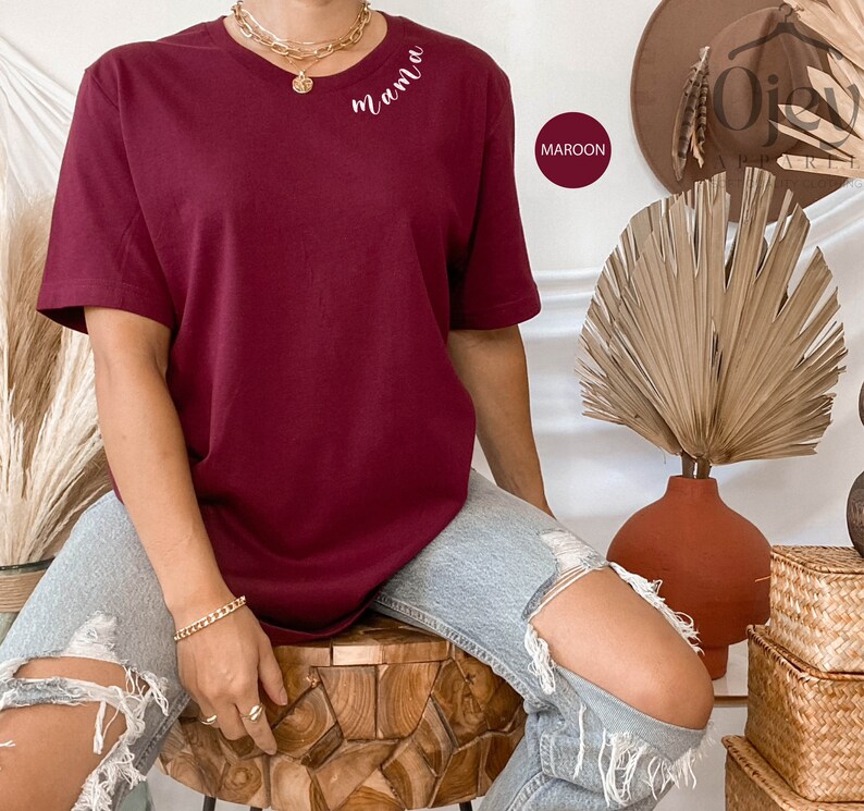 K&ouml;nnte beinhalten: Ein maroonfarbenes T-Shirt mit dem Wort "mama" auf der Vorderseite gestickt. Das Shirt wird von einer Person getragen, die auf einem Holzhocker sitzt.
