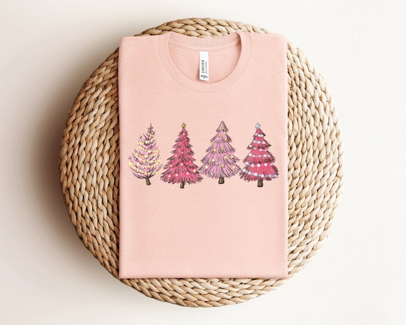 May include: A pink t-shirt featuring a design of four pink Christmas trees with gold star and light accents.