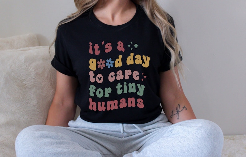 May include: A black t-shirt with the text "it's a good day to care for tiny humans" in colorful retro-style lettering.