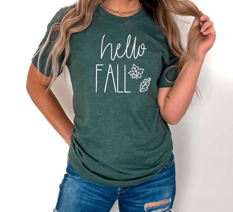May include: A dark green t-shirt with the text "hello fall" in white lettering. The shirt features a white outline of a maple leaf and a small leaf.
