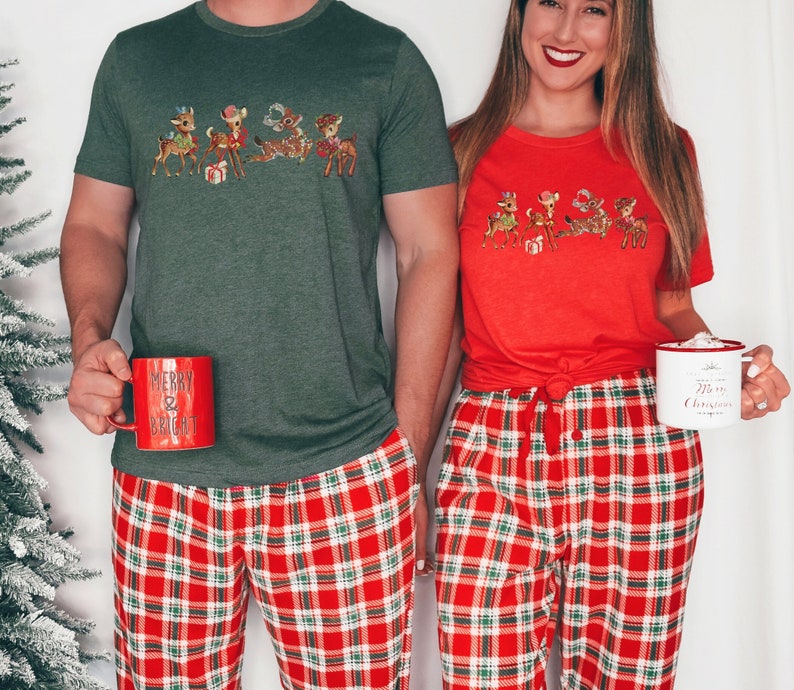 Pu&ograve; includere: Una coppia indossa pigiami natalizi abbinati con un design a renna. L'uomo indossa una camicia verde con pantaloni da pigiama a quadri rossi e verdi. La donna indossa una camicia rossa con pantaloni da pigiama a quadri rossi e verdi. Entrambi tengono in mano delle tazze.