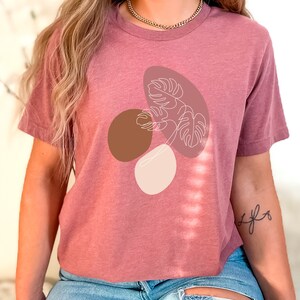 Puede incluir: Una camiseta malva con un dibujo de l&iacute;nea blanca de una hoja tropical con formas abstractas marrones y beige.