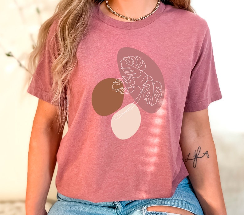 Puede incluir: Una camiseta malva con un dibujo de l&iacute;nea blanca de una hoja tropical con formas abstractas marrones y beige.