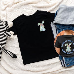 Puede incluir: Dos camisetas negras con un gr&aacute;fico de conejo colorido. Una camiseta tiene el texto "Me encanta cuando me llamas Hoppa" en la espalda.