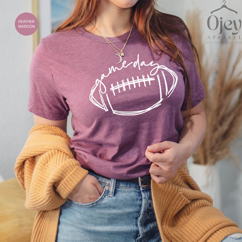 Peut inclure: Un t-shirt en marron chin&eacute; avec un motif blanc de football et le texte "game day".
