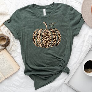 May include: A green t-shirt with a leopard print pumpkin design. The shirt is on a white surface with a book, a clock, a coffee cup, and a hat.