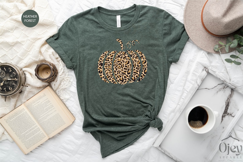 May include: A green t-shirt with a leopard print pumpkin design. The shirt is on a white surface with a book, a clock, a coffee cup, and a hat.