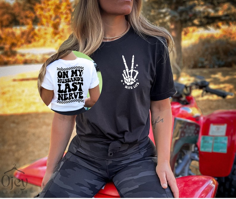 May include: A black t-shirt with a white skeleton hand making a peace sign and the text "Wife Life". A white t-shirt with the text "On My Husband's Last Nerve" is visible in the background.