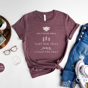 K&ouml;nnte beinhalten: Ein T-Shirt in Heather-Maroon mit einem wei&szlig;en Grafikdesign, das eine Biene, B&auml;ume und Wellen zeigt. Der Text auf dem Shirt lautet "Help More Bees, Plant More Trees, Clean The Seas".