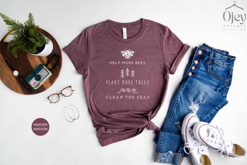 K&ouml;nnte beinhalten: Ein T-Shirt in Heather-Maroon mit einem wei&szlig;en Grafikdesign, das eine Biene, B&auml;ume und Wellen zeigt. Der Text auf dem Shirt lautet "Help More Bees, Plant More Trees, Clean The Seas".
