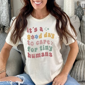 May include: A white t-shirt with the text "it's a good day to care for tiny humans" in colorful retro-style lettering with flowers and hearts.