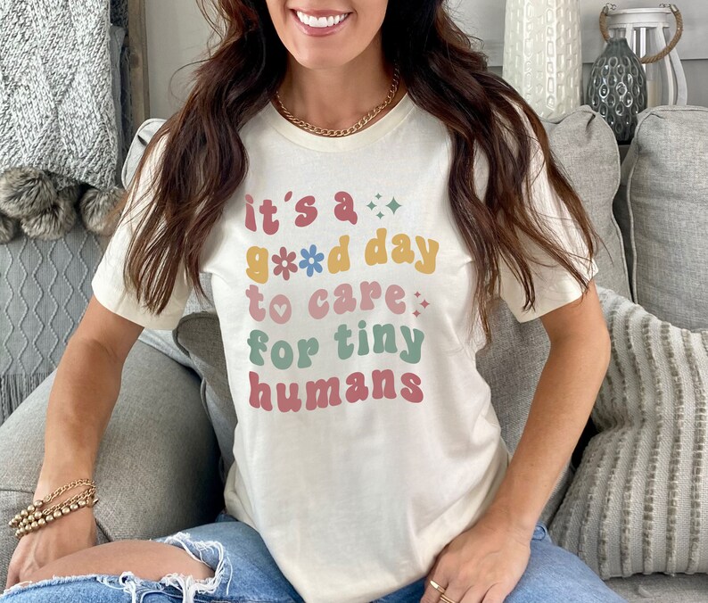 May include: A white t-shirt with the text "it's a good day to care for tiny humans" in colorful retro-style lettering with flowers and hearts.
