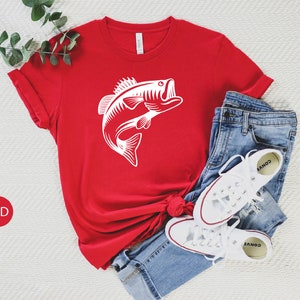 May include: Red t-shirt featuring a white graphic of a bass fish leaping out of the water. The shirt is laid out on a white surface with a pair of white trainers and blue jeans.